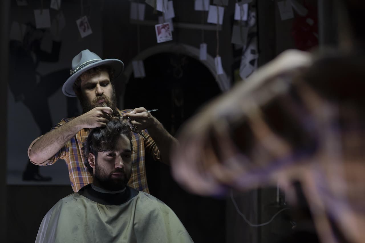 A barber in a felt hat cutting a seated customer's hair with scissors, seen in the mirror